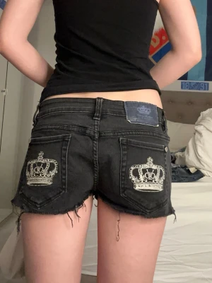 Svarta lågmidjade jeansshorts med krona - Säljer ett par svarta lågmidjade jeansshorts från victoria Beckham med broderade vita kronor på bakfickorna. 40cm raktöver midjan❤️