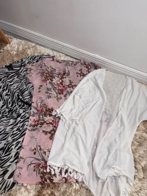 3 strandklänningar i olika mönster - Säljer tre strandklänningar: en med zebramönster i svartvitt, en rosa med blommigt tryck och en vit transparent kimono med fransar. Perfekta att slänga över bikinin på stranden. Alla är i tunt, luftigt material.
