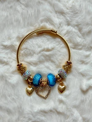 Guldarmband med blå pärlor och hjärtan - Snyggt armband i guldfärgad metall med glittrande blå och vita pärlor. Armbandet har tre hängande hjärtberlocker, varav ett är dekorerat med små stenar. Perfekt accessoar för att lyfta din outfit med lite extra bling.