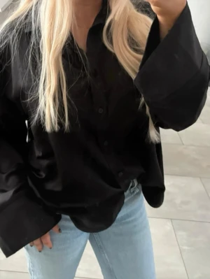 Svart oversized skjorta med knappar satin  - Säljer en svart oversized skjorta med klassisk krage och knappar framtill. Skjortan har långa, vida ärmar och är tillverkad i ett blankt material som ger en snygg look. Perfekt att styla med jeans eller kjol.