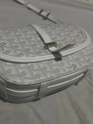 Vit axelväska - Snygg vit axelväska med det klassiska mönstret och justerbar axelrem. Väskan har ett lock med spänne i silverfärg och är gjord i tåligt canvasmaterial med läderdetaljer. Perfekt för dig som vill ha en unik och trendig accessoar.