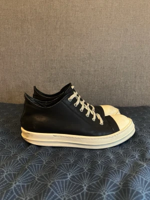 Svarta Rick Owens sneakers i läder - Säljer ett par svarta sneakers från Rick Owens med vita snören och vit tå samt sula. Skorna är låga och har en chunky siluett med en desstresresed och klassisk design. Tillverkade i läder för en lyxig känsla och edgy look. Rätt så använda kommer utan låda 