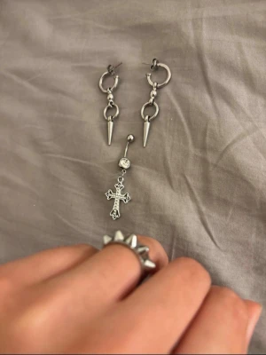 Smyckeset med spikes och kors - Säljer ett set med silvriga smycken: ett par örhängen med ringar och hängande spikes, en navelpiercing med glittrande sten och kors, samt en ring med nitar. Perfekt för dig som gillar alternativ stil och detaljer i metall.