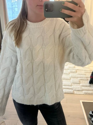 Vit kabelstickad tröja - Säljer en vit kabelstickad tröja med rund hals och långa ärmar. Tröjan har ett klassiskt flätmönster och ribbade muddar vid ärmslut och nederkant. Perfekt för kalla dagar och enkel att matcha med jeans eller kjol. Den är använd en del och därför lite nopprig. Tröjan är också liten i storleken, för mig som annars brukar ha  M.