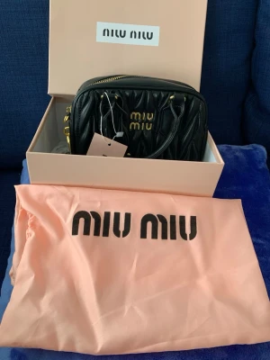 Svart handväska från Miu Miu - Snygg svart handväska från Miu Miu i quiltat läder med guldfärgade detaljer och logotyp framtill. Väskan har ett topphandtag och kommer med original dustbag och box. Perfekt för dig som gillar lyxiga accessoarer med exklusiv känsla. Inköpt utomlands som present har ej kvitto el certifikat alltså inga garantier ang märket. Har pga detta satt ett lågt pris. Vid frågor skriv!