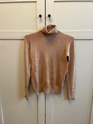 Beige polotröja i mjukt stickat material - Säljer en beige stickad polotröja med lång ärm och ribbade muddar. Tröjan har en hög polokrage och är tillverkad i ett tunt, mjukt material som känns skönt mot huden. Perfekt för lager på lager och enkel att matcha med olika outfits.