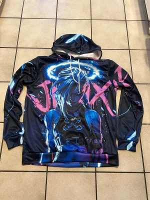 Jinx hoodie med neontryck storlek L - Snygg hoodie med stor färgstark Jinx-illustration från League of Legends i neonblått, rosa och svart. Hoodien har huva med vita snören och tryck över hela plagget. Perfekt för dig som gillar gaming och streetwear.
