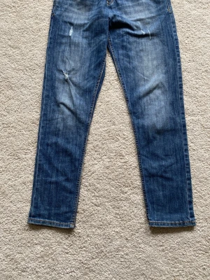 Blå raka jeans med slitningar - Säljer ett par klassiska blå jeans med raka ben och snygga slitningar på framsidan. I jättebra skick Jeansen har fem fickor och kontrastsömmar. Perfekta för en avslappnad och trendig look. 