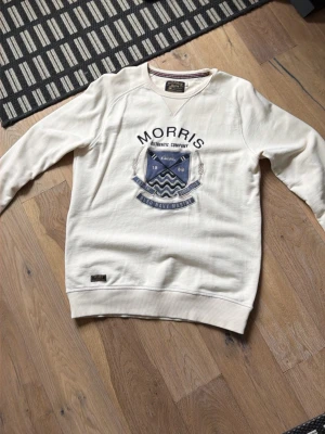 Vit sweatshirt från Morris, S - Vit sweatshirt från Morris med tryck i blått och grått på bröstet. Tröjan har rund halsringning med randig detalj på insidan och ribbade muddar vid ärmslut och nederkant. Perfekt för en avslappnad och stilren look.