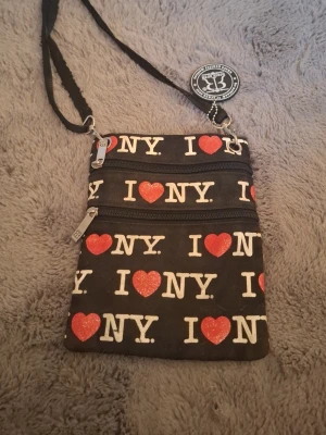 I Love NY svart axelväska - Snygg svart axelväska med texten 'I ♥ NY' i vitt och rött tryck över hela väskan. Väskan har två dragkedjefack framtill och justerbar axelrem. Perfekt för dig som gillar streetstyle och vill ha en unik accessoar med New York-vibe.