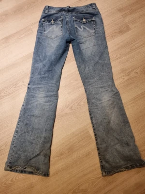 Blå bootcut jeans med fickdetaljer - Snygga blå bootcut jeans med dekorativa fickor bak med knapp och sömmar i zigzag. Perfekta för en avslappnad och trendig look. Pris kan diskuteras. 