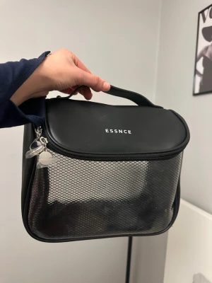 Svart necessär från ESSNCE - Snygg svart necessär från ESSNCE med genomskinlig mesh på framsidan och dragkedja med silverfärgade detaljer. Praktiskt handtag upptill och rymligt fack för smink eller småprylar. Perfekt för att hålla ordning på dina grejer.