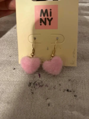 Rosa fluffiga hjärtörhängen MiNY - Supersöta örhängen från MiNY med fluffiga rosa hjärtan. Hängena har guldfärgade krokar och hjärtana är täckta av mjukt, lurvigt material som ger en gullig och lekfull look. Perfekta för dig som vill sticka ut med något unikt och charmigt.