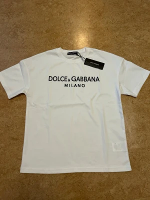 Vit Dolce & Gabbana t-shirt - Snygg vit t-shirt från Dolce & Gabbana med svart logotyp och Milano-tryck på bröstet. Klassisk rund hals och korta ärmar. Tillverkad i mjuk bomull. Ny med lappen kvar. Storlek xs, passar någon med storlek small, den är oversize