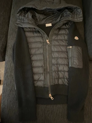 Svart cardigan från Moncler - Svart cardigan från Moncler med quiltad front i dun och stickade ärmar. Jackan har huva, dragkedja framtill och en ficka med dragkedja på ärmen. Moncler-logga på armen ger en exklusiv vibe. Perfekt för dig som gillar streetstyle och premiumkänsla.