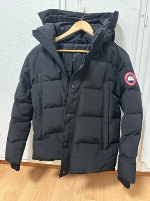 Svart dunjacka från Canada Goose - Snygg svart dunjacka från Canada Goose med klassisk patch på ärmen. Jackan har huva, dragkedja och tryckknappar framtill samt flera praktiska fickor. Perfekt för kalla vinterdagar och riktigt varm tack vare dunfyllningen.