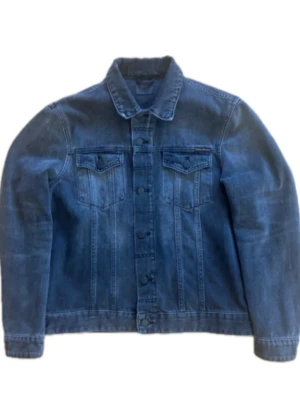 Grå jeansjacka från Nudie Jeans - Klassisk grå jeansjacka från Nudie Jeans med raka linjer, två bröstfickor med lock och knappar framtill. Jackan har en snygg tvättad finish och diskret logga på bröstet. Perfekt för dig som gillar tidlös streetstil och denim. OBS. Tveka inte på att höra av er vid frågor!