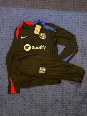FC Barcelona träningsset Nike svart - Säljer ett svart träningsset från Nike och FC Barcelona med långärmad tröja och byxor. Setet har röda och blå detaljer, klubbmärke och Spotify-logga på bröstet samt Nike.