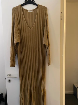 Beige ribbad långklänning från Rodebjer - Långklänning från Rodebjer i beige med ribbad struktur och v-ringning. Klänningen har lång ärm och en loose passform som ger en avslappnad och trendig look. Perfekt för dig som gillar stilrena och bekväma plagg.