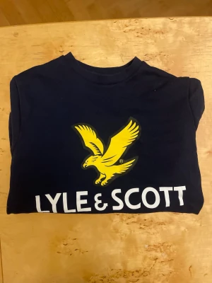 Marinblå Lyle & Scott tjocktröja  - Snygg marinblå t-shirt från Lyle & Scott med stor gul logga och text framtill. Klassisk rund halsringning och korta ärmar. Perfekt för dig som gillar stilrena plagg med tydlig branding.