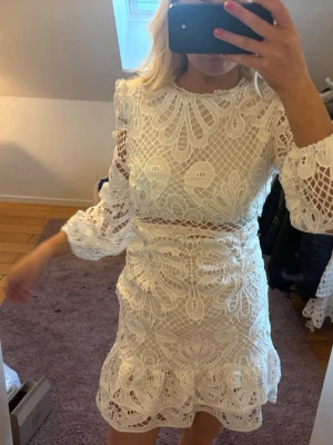 Vit spetsklänning med volang - Supersnygg vit kort klänning i spets med volangdetaljer och långärmade ärmar. Klänningen har ett genombrutet mönster, rund halsringning och markerad midja. Perfekt för dig som vill sticka ut med en romantisk look.