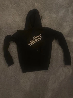 Svart Saint Laurent hoodie - Säljer en svart hoodie från Saint Laurent med stor vit logga på bröstet. Klassisk huva med dragsko och känguruficka framtill. Storlek S. Tillverkad i mjukt bomullsmaterial som är skönt att bära. Perfekt för dig som gillar streetwear och exklusiva märken. Priset är ej hugget i sten!🤗