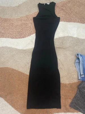 Svart långklänning - En stilren svart långklänning med ärmlös design. Klänningen har en figurnära passform och är tillverkad i ett mjukt, stretchigt material som ger en smickrande siluett. Perfekt för dig som gillar minimalistisk och elegant stil.