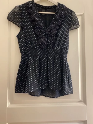 Mörkblå prickig blus med volanger - Söt mörkblå blus med vita prickar och volangdetaljer längs v-ringningen. Blusen har korta ärmar i transparent material och smock i midjan för en feminin siluett. Perfekt till jeans eller kjol för en trendig look.💞säljer pga den inte kommer till användning💞