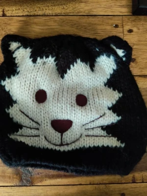 Svart stickad mössa med kattmotiv - Stickad svart mössa med ett stort vitt kattansikte framtill, inklusive broderade morrhår och öron som sticker ut upptill. Mössan har en mjuk och stretchig passform och är perfekt för dig som vill ha en lekfull accessoar till hösten eller vintern.