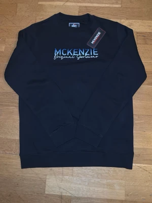 Svart sweatshirt från McKenzie - Svart sweatshirt från McKenzie med blått och vitt broderat tryck på bröstet där det står 'McKenzie Original Sportswear'. Klassisk rund halsringning och långa ärmar. Perfekt för en avslappnad streetwear-look.