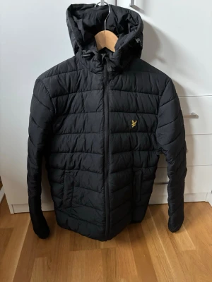 Lyle & scott jacka - Snygg svart pufferjacka från Lyle & Scott med gul logga på bröstet. Jackan har huva, dragkedja framtill och två sidofickor. Quiltad design med horisontella sömmar och en modern passform. Perfekt för kalla vinterdagar.