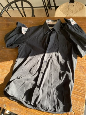 Svart skjorta från Burberry - Klassisk svart skjorta från Burberry med ikoniskt rutigt mönster på insidan av kragen och ärmsluten. Skjortan har knappar framtill, bröstficka och är tillverkad i mjuk bomull. Perfekt för dig som gillar stilrena plagg med exklusiva detaljer.