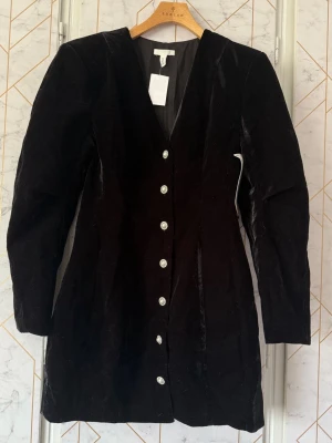 Provkollektion svart sammetsklänning med knappar - Stilren svart Sasha sammetsklänning med v-ringning och långa ärmar. Klänningen har dekorativa silverfärgade knappar framtill och en figursydd passform som ger en elegant siluett. I material recycle polyester. Denna är troligtvis från en provkollektion som har hängt ute på en mässa