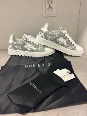  Numeris - Snygga sneakers från Numeris i grått och vitt camomönster. Skorna har tjock vit sula med räfflad undersida, vita skosnören och tåparti i vitt. Tillverkade i textil och syntetmaterial. Perfekta för dig som vill sticka ut med en unik look. Diskuterat pris gärna. 