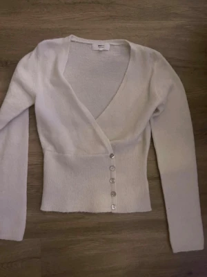 Vit omlottkofta med knappar - Säljer en vit omlottkofta med långa ärmar och sex silvriga knappar framtill. Koftan har en djup v-ringning och är ribbstickad i midjan för en snygg passform. Perfekt att styla över ett linne eller topp.