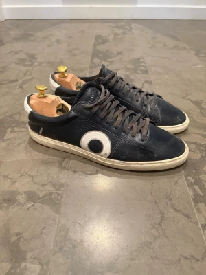 Oliver Cabell - Stilrena och tidlösa Oliver Cabell Low 1 sneakers i mörkblått läder. En premium, minimalistisk design som passar perfekt till både vardag och mer uppklädda outfits.  Tillverkade i högkvalitativt läder med en ren siluett och signaturdetalj på sidan. Skorna är använda och har naturliga veck i lädret samt mindre tecken på användning, men är fortfarande i bra skick med mycket liv kvar.  Perfekta att bära till jeans, chinos eller mer dressade looks.  ✔ Märke: Oliver Cabell low 1 ✔ Strl: 46