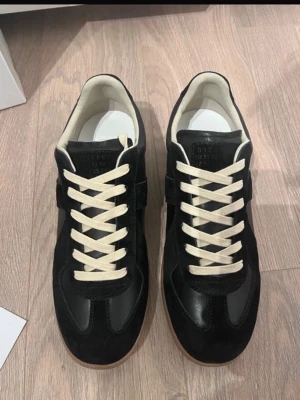  Maison Margiela gats - Maison Margiela Replica sneakers i svart mocka och läder med vita kontrasterande snören och vit innersula. Klassisk låg modell med diskret logotyp på plösen och gummisula. Perfekta för dig som gillar stilrena och tidlösa sneakers.