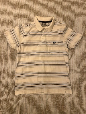 Randig vit piké från Nike - Vit pikétröja från Nike med tunna blå och svarta ränder. Bra skick, inga hål. Jag är 174 och den sitter bra på mig.