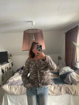 Brunmelerad kofta med knappar - Säljer en brunmelerad kofta med grovstickad struktur och raka ärmar. Koftan har rund halsringning och knäpps framtill med dekorativa knappar. 