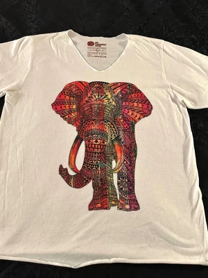 Vit t-shirt med färgglad elefant L - Säljer en vit t-shirt med v-ringning och ett stort, färgglatt elefanttryck på bröstet. Trycket har detaljer i rött, orange, gult och rosa med ett unikt, mönstrat motiv. Perfekt för dig som vill sticka ut med en cool och konstnärlig look., bomull 