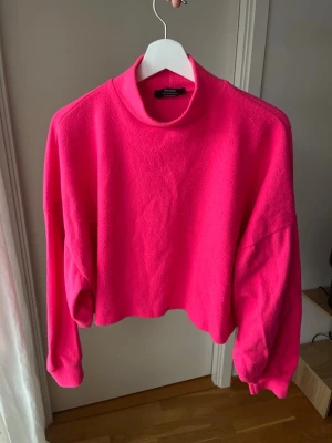 Rosa fleece-tröja - Säljer en chockrosa croppad fleece-tröja från Bershka. Tröjan har hög krage, avslappnad passform och långa ärmar med breda muddar. Perfekt för dig som vill sticka ut med färg och gillar mjuka material.