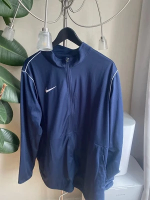 Marinblå Nike träningsjacka - Snygg marinblå träningsjacka från Nike med vit logga på bröstet och vita detaljer längs axlarna. Jackan har halv dragkedja framtill, hög krage och är tillverkad i ett mjukt funktionsmaterial som passar perfekt till träning eller uppvärmning.