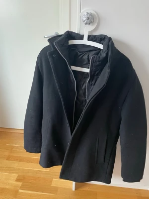 Vår/vinter rock. - Stilren svart rock med avtagbar ”innejacka”. Rocken har två sidofickor och quiltat innerfoder för extra värme. Passar perfekt till höst och vinter när du vill hålla stilen enkel och snygg. Materialet ser ut att vara ull.                                        XS men passar S, använd 3 gånger. Köpt för strax under 2000kr