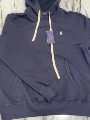 Ralph lauren hoodie  - Mörkblå hoodie från Polo Ralph Lauren med vit broderad logga på bröstet och vita dragsnören i huvan. Klassisk känguruficka framtill och ribbade muddar vid ärmar och nederkant. Tillverkad i mjuk bomullsblandning för skön känsla.