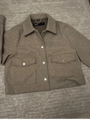 Jacka - Stilren beige overshirt från Zara med klassisk krage och stora bröstfickor med lock. Knäpps med silvriga knappar framtill. Overshirten har en rak passform och är gjord i ett mjukt, ull-liknande material som ger en trendig look.