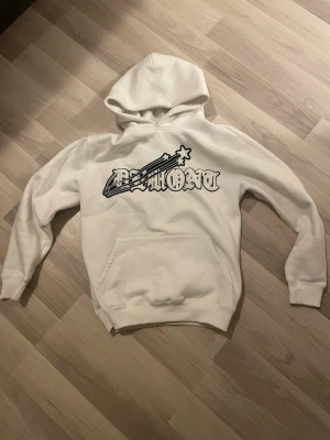 Vit hoodie från Demont med tryck - Säljer en vit hoodie från Demont i storlek S. Hoodien har ett stort svart tryck med text och stjärndesign på bröstet samt en klassisk känguruficka framtill. Tillverkad i mjukt material och har en rymlig huva. Perfekt för en avslappnad streetwear-look.