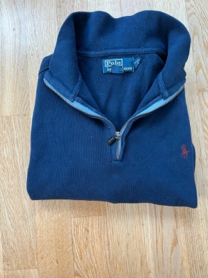 Mörkblå halvzip-tröja från Polo - Mörkblå långärmad tröja från Polo Ralph Lauren med halvzip och klassisk krage. Tröjan har ett diskret rött broderat logomärke på bröstet och är tillverkad i mjukt bomullsmaterial. Perfekt för en avslappnad och stilren look.