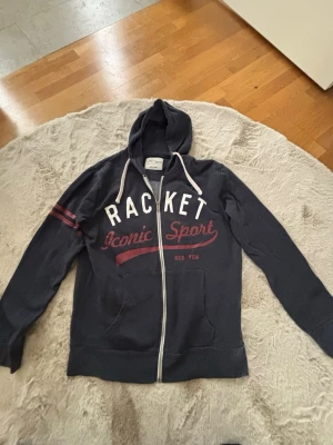 Mörkblå hoodie med dragkedja - Mörkblå hoodie med vit dragkedja och huva. Tryck i vitt och rött framtill med texten 'RACKET Iconic Sport'. Två röda ränder på ena ärmen och vita dragsnören i huvan. Perfekt för en sportig och avslappnad stil.