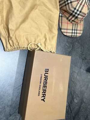 Burberry beige rutig keps - Säljer en klassisk Burberry keps i beige med det ikoniska rutmönstret i svart, vitt och rött. Kepsen har böjd skärm och justerbar rem bak. Tillverkad i bomull och levereras med originalpåse och box. Perfekt accessoar för en trendig streetwear-look. Säljes billig behöver pengar snabbt 