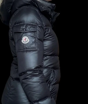 Svart dunjacka från Moncler - Snygg svart dunjacka från Moncler med glansig finish, huva och tvåvägsdragkedja. Jackan har fickor med dragkedja framtill och en ficka med lock på ärmen där Moncler-loggan sitter. Perfekt för kalla vinterdagar.